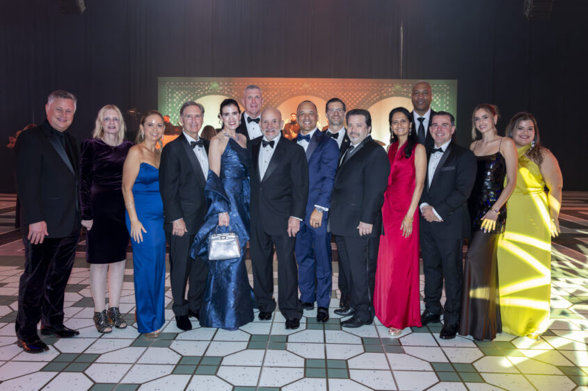 A group of fifteen people dressed in formal evening wear poses together on a patterned floor, smiling at the camera against a decorative backdrop with green and gold lighting, truly embodying the spirit of Golden Angels and making a Real Impact. Lifestyle