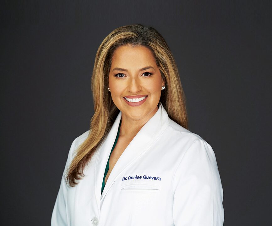 A smiling woman with long light brown hair wears a white lab coat embroidered with "Dr. Denise Guevara," a dermatologist who debunks Spring skin myths, and stands against a dark gray background. Lifestyle