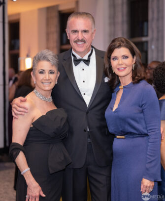 Three elegantly dressed adults pose together at a formal event, exuding the grace of Golden Angels. The woman on the left wears an off-shoulder black dress, the man in a tuxedo, and the woman on the right shines in a long-sleeve blue dress. Lifestyle