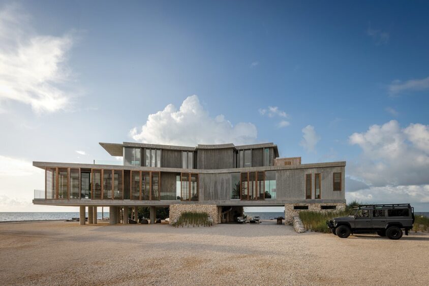 A modern, multi-level house designed by Max Strang with large glass windows and wooden accents stands on a Florida beach, elevated on pillars. A dark SUV is parked on the gravel driveway under a partly cloudy sky. Lifestyle