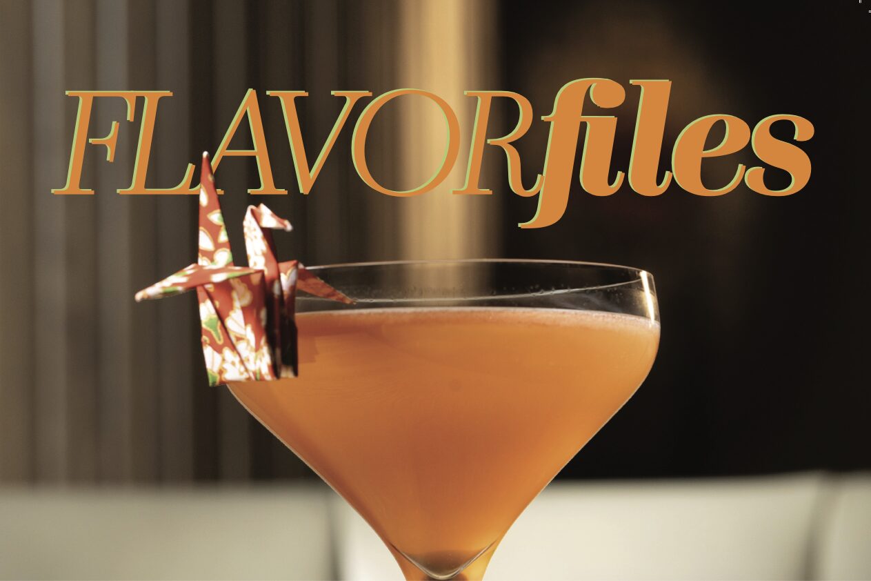 A cocktail in a martini glass garnished with an origami crane, with "FLAVORfiles" in bold orange letters and "April Flavor Files" subtly featured above. The background is softly blurred. Lifestyle