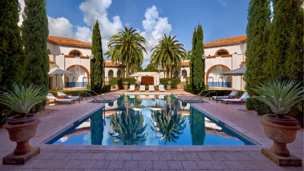 A luxurious outdoor pool at the Boca Hotel is surrounded by lounge chairs, tall trees, palm trees, and Mediterranean-style buildings with arches and red-tiled roofs under a partly cloudy sky. Lifestyle