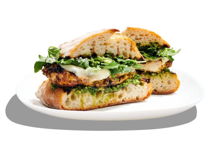 A Clean By Design sandwich on ciabatta bread filled with grilled chicken, melted cheese, fresh greens, and pesto sauce, served on a white plate against a white background. Lifestyle
