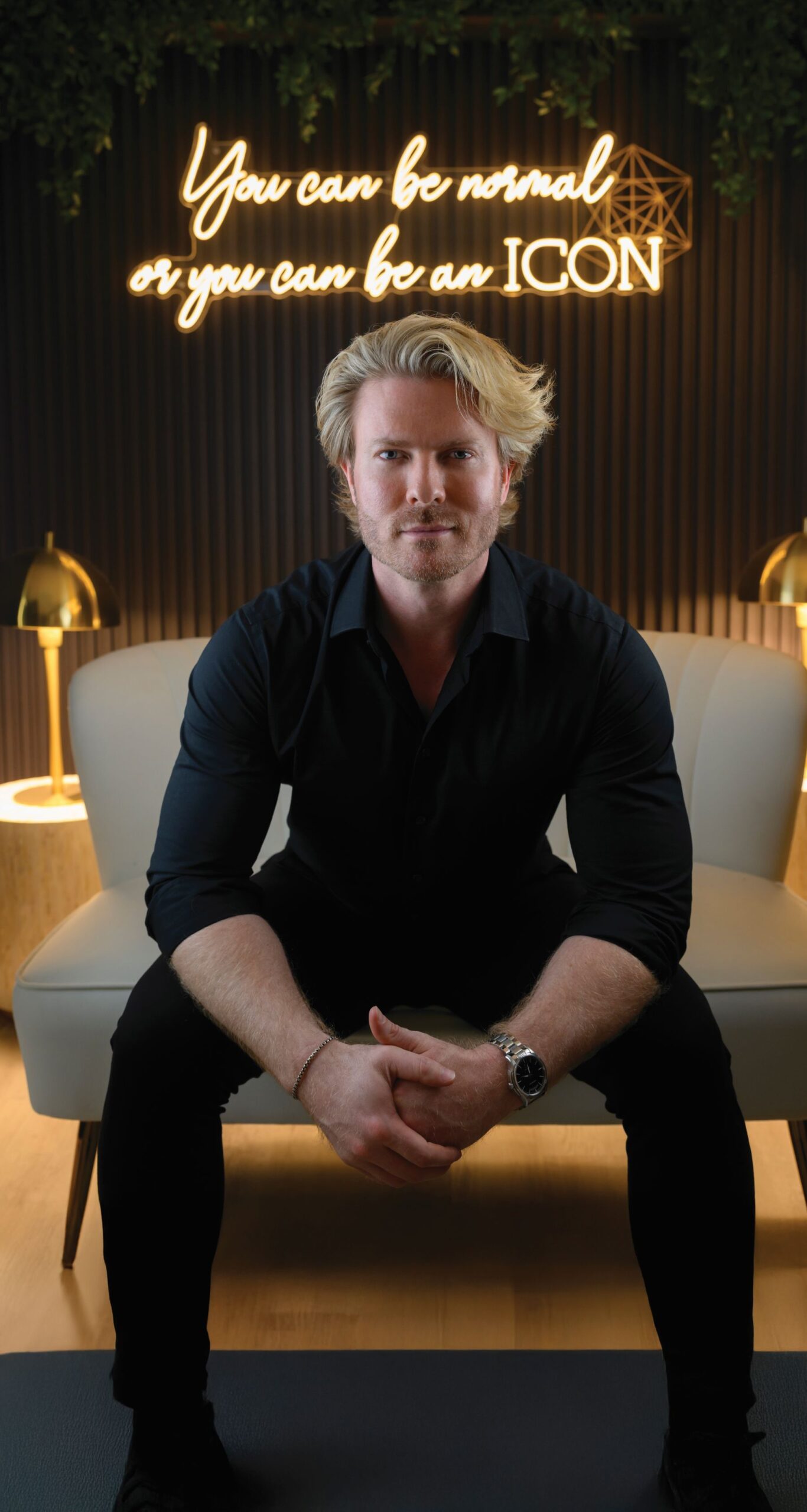 A hot & unbothered man with blond hair and a black shirt sits on a light-colored couch, hands clasped, in a modern room. Behind him, a neon sign boldly declares, "You can be normal or you can be an ICON. Lifestyle