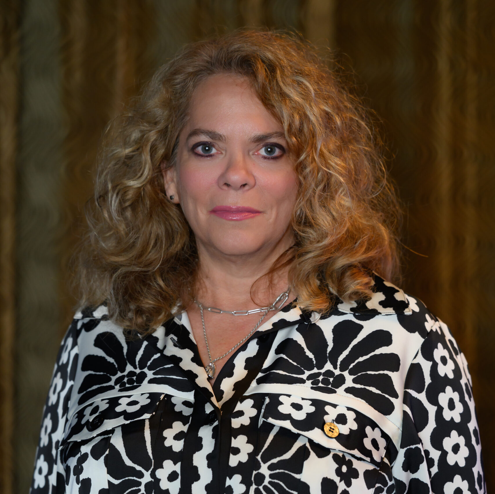 A woman with curly blonde hair, wearing a black-and-white floral patterned blouse and a belt, stands in front of a gold curtain, looking directly at the camera with a neutral expression—ready to lead her team. Lifestyle
