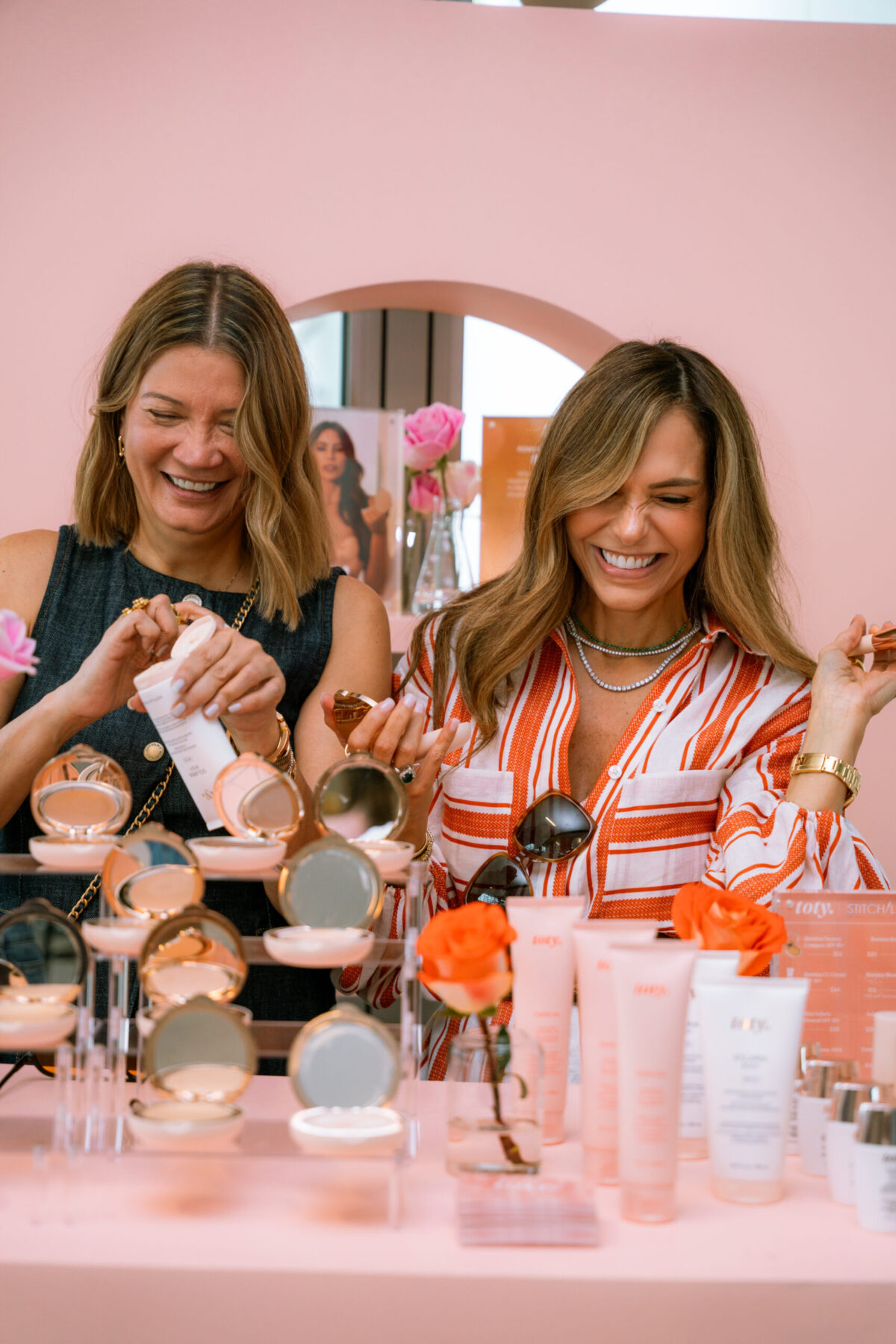 Two women smiling and laughing while testing skincare and makeup products at a Stitch Lab Pop-Up display table decorated with orange roses and pink packaging. The pastel pink background creates a cheerful atmosphere at W Hotel Fort Lauderdale. Lifestyle