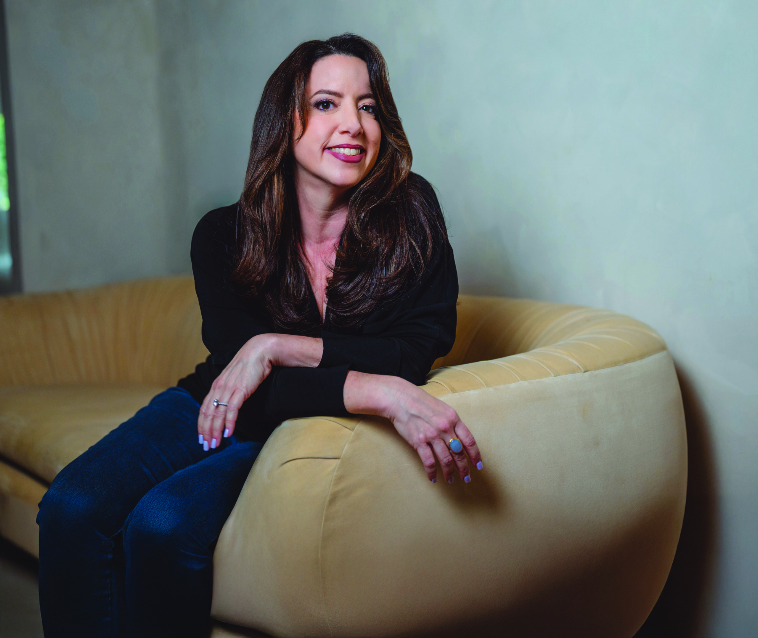 A woman with long brown hair, wearing a black top and blue jeans, sits smiling on a light yellow curved sofa against a pale wall—her calm presence belying any sense of being Shaken to the Core. Lifestyle