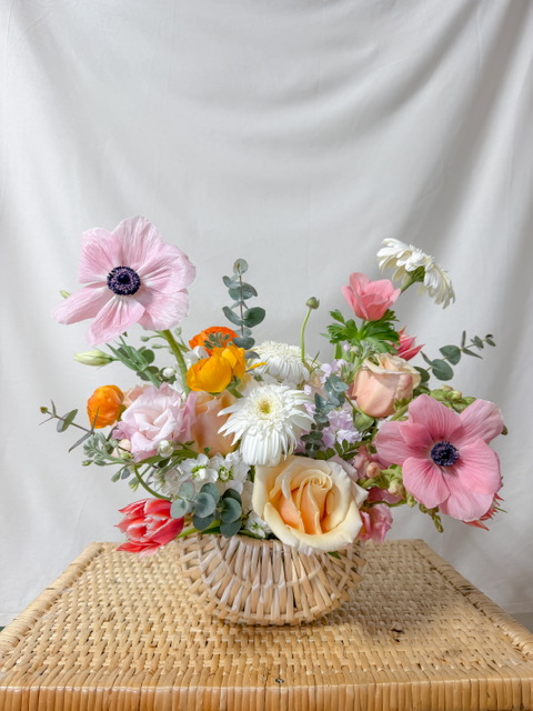 A woven basket vase holds a colorful arrangement of flowers in full bloom, including pink anemones, white daisies, orange ranunculus, and pale roses, set against a light fabric backdrop on a wicker surface. Lifestyle