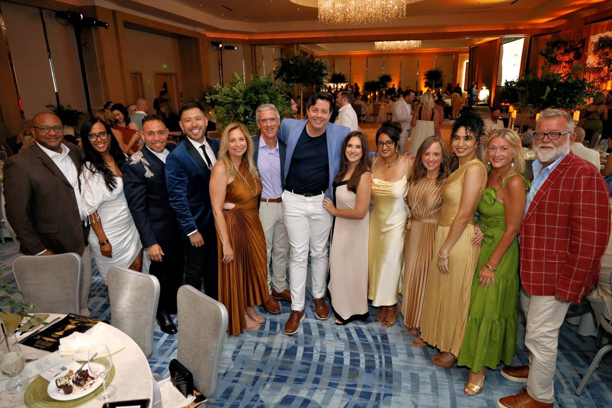 A group of twelve adults in semi-formal attire smile for a photo at the Golden Hour Gala, an elegant banquet event hosted by Broward Partnership to combat homelessness, with round tables and chandeliers adorning the room. Lifestyle