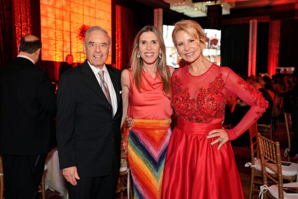 Three adults pose and smile at a formal United Way event with red and orange lighting. The man wears a dark suit and tie, while the two women wear colorful dresses—one with a multicolored skirt and the other in a long red gown. Lifestyle