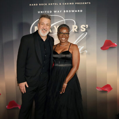A man in a black suit and a woman in a black gown smile together at the United Way Broward Mayor's Gala, standing before a backdrop adorned with red rose petals, celebrating their support for United Way’s mission. Lifestyle