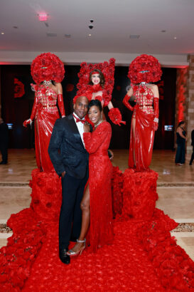 A couple poses on a red rose-covered walkway, with three performers in dramatic red dresses and elaborate rose headpieces standing behind them at a glamorous United Way event in a formal setting. Lifestyle