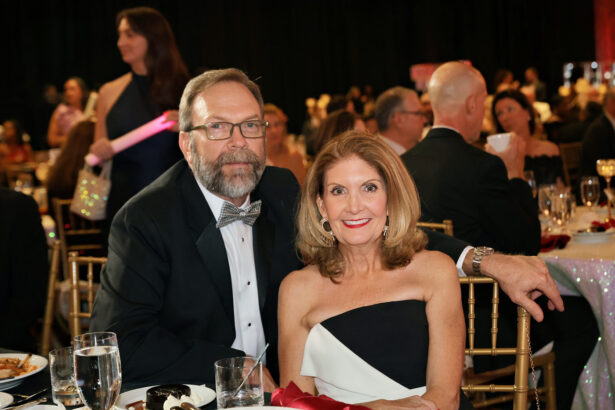 A man in a suit and bow tie sits beside a woman in a black-and-white strapless dress at a United Way formal event, surrounded by other guests at decorated tables. Both are smiling at the camera. Lifestyle