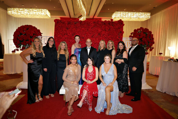 A group of people in formal attire pose for a photo at a United Way event in a decorated ballroom, with a red rose floral backdrop and chandeliers above. Some are seated in front, while others stand behind them, all smiling at the camera. Lifestyle