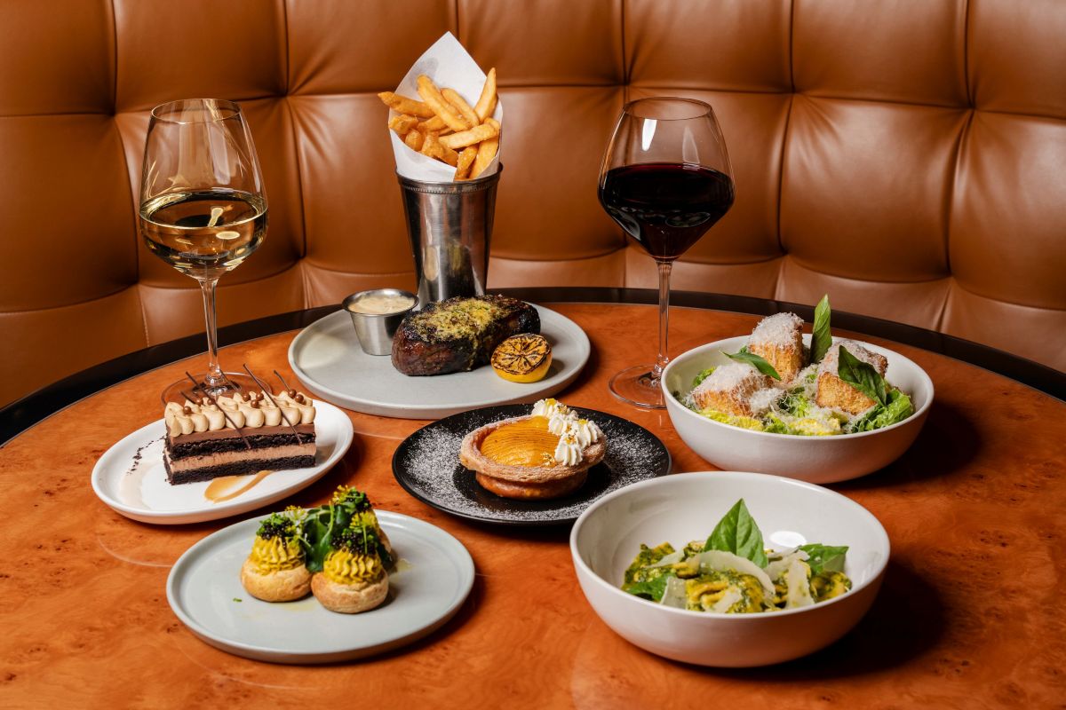 A table set with two glasses of wine, steak with fries—featuring some of the best steaks in Miami—a salad, pasta, deviled eggs, chocolate dessert, and a tart, all atop a wood table with a tufted leather booth in the background. Lifestyle