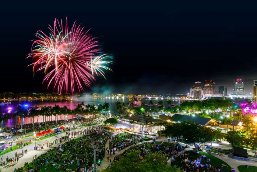 A vibrant fireworks display lights up the night sky over a lively city waterfront on Independence Day, with crowds gathered in a park, colorful lights reflecting on water, and tall buildings in the background. Lifestyle