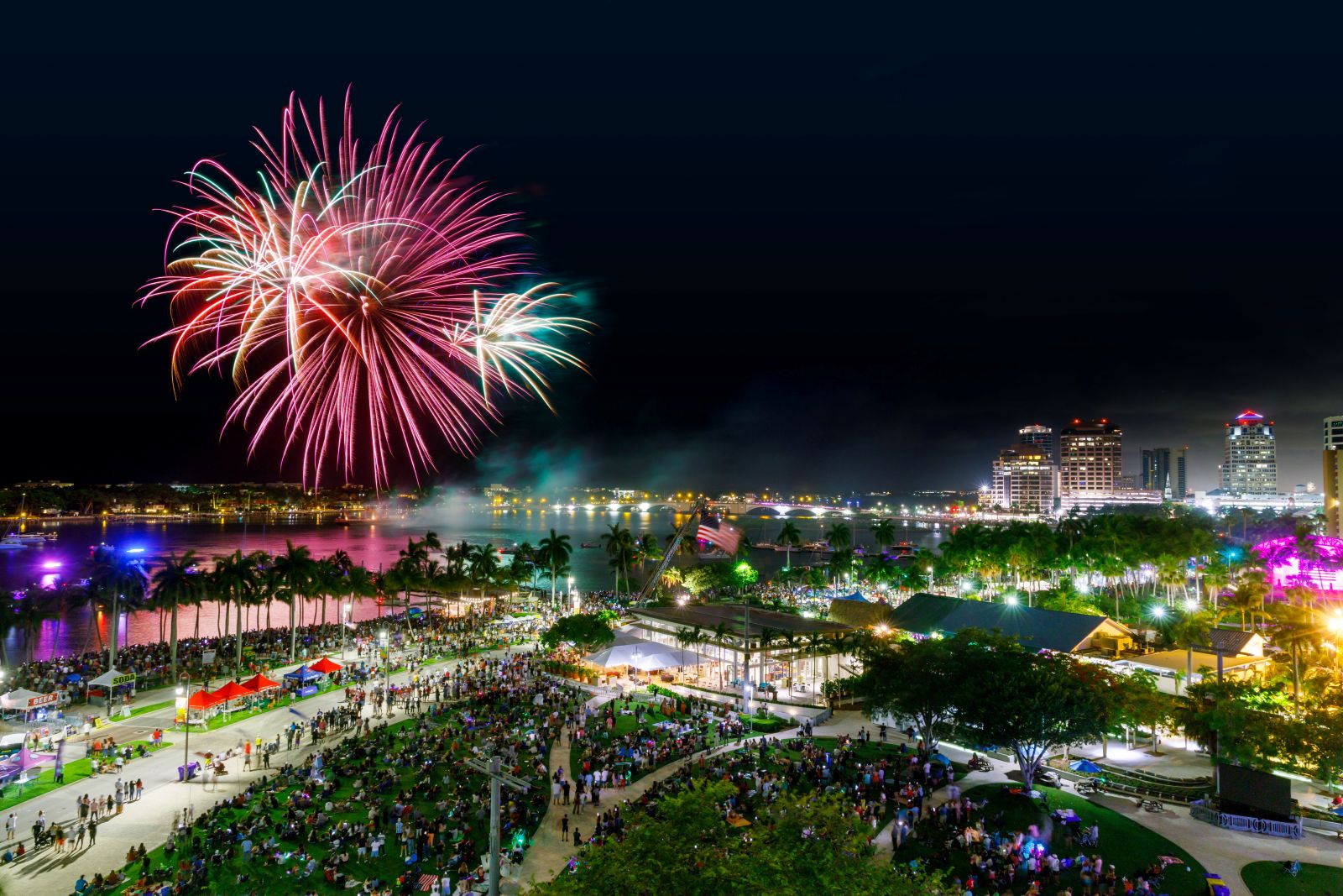 A vibrant fireworks display lights up the night sky over a lively city waterfront on Independence Day, with crowds gathered in a park, colorful lights reflecting on water, and tall buildings in the background. Lifestyle