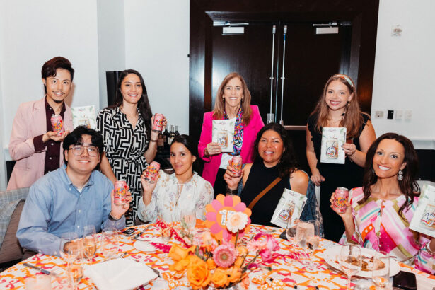 A group of eight people sit and stand around a decorated table, smiling and holding colorful canned drinks and pouches in a bright, festive setting. The table has pink and orange floral centerpieces as the party Gets Groovy with Children’s Opportunity Group. Lifestyle