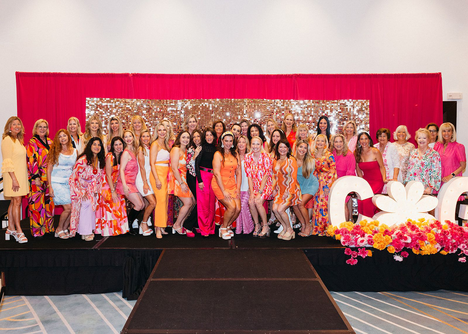 A large group of women in colorful, vibrant dresses poses together on a stage with a sparkling gold backdrop, pink and red curtains, and floral decorations as the Children’s Opportunity Group Gets Groovy in front of large white letters and a daisy. Lifestyle