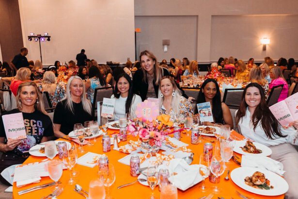 A group of seven women from the Children’s Opportunity Group sit around a table with orange and white decor at a formal event, smiling and holding up books. Plates of food, drinks, and floral centerpieces add to the Gets Groovy atmosphere. Lifestyle