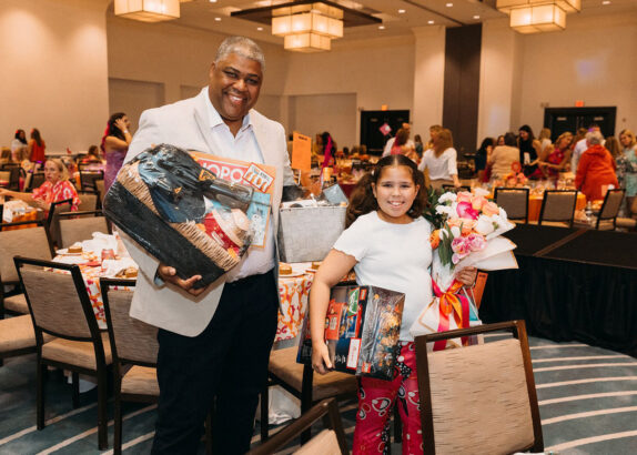 A smiling man and a young girl stand together in a brightly lit event hall, holding gift baskets, flowers, and board games at the Children’s Opportunity Group Gets Groovy event. People are seated at round tables in the background. Lifestyle