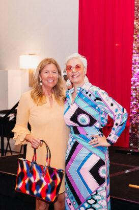 Two women stand together smiling at the Gets Groovy event. One wears a light yellow dress with a colorful bag; the other sports a vibrant geometric-patterned dress with red-tinted glasses. A red curtain and shiny backdrop are behind them. Lifestyle