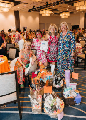 Four women in colorful dresses pose together at the Children’s Opportunity Group Gets Groovy indoor event, smiling amid gift baskets filled with various items. Other attendees can be seen in the background at decorated tables. Lifestyle