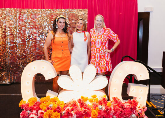 Three women in retro-inspired dresses stand on a stage decorated with pink and orange flowers and large illuminated letters "C" and "G" for the Children’s Opportunity Group Gets Groovy event, set against a sequin backdrop and vibrant pink curtains. Lifestyle