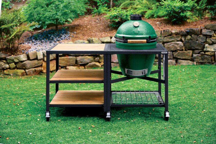 A green ceramic outdoor grill is set in a black metal cart with wooden shelves, showing that it's easy being green amid the grass, stone wall, and lush garden backdrop. Lifestyle