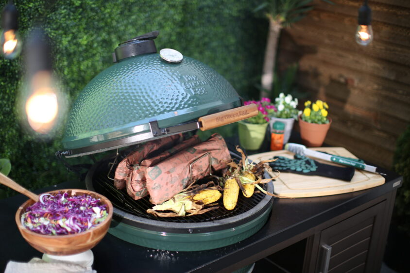 A sustainable green ceramic grill with wrapped meat and corn on the cob inside, beside a bowl of salad, kitchen tools, potted flowers, and a leafy green backdrop with hanging lights. Lifestyle