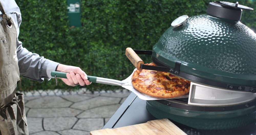 A person uses a metal pizza peel to remove a cooked pizza from a green ceramic outdoor grill, proving it’s easy being green, with a wooden cutting board on the side and lush greenery in the background. Lifestyle