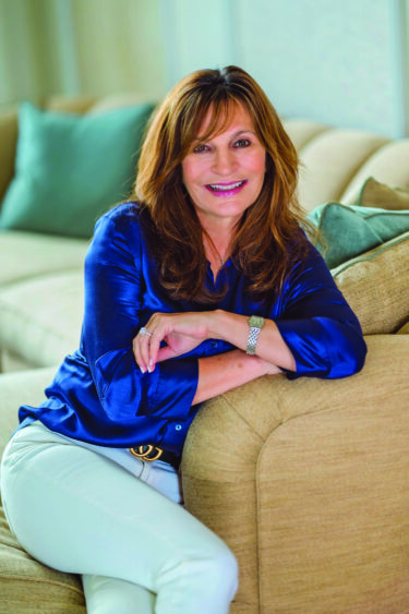 A woman with long brown hair, wearing a shiny blue blouse and white pants, sits smiling on a beige sofa with her arm resting on the couch back, embodying audience engagement. Neutral-colored pillows are in the background. Lifestyle