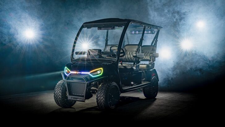 A black, modern golf cart with colorful LED lights on the front is parked indoors, surrounded by dramatic fog and bright spotlights in the background. Lifestyle
