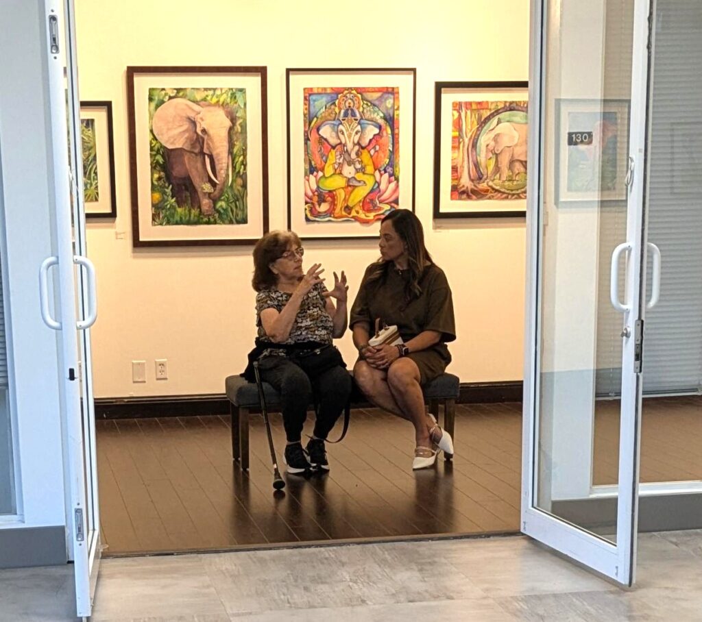 ArtServe, engaged in conversation. Behind them are colorful paintings of animals, including an elephant and an elephant-headed figure. The gallery doors are open, inviting viewers to explore the art of language and creativity inside. Lifestyle
