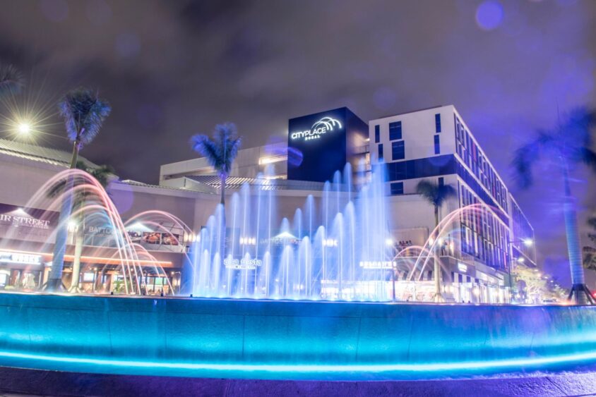 A night view of CityPlace Doral with palm trees and a brightly lit fountain featuring blue and pink lights, surrounded by modern buildings under a cloudy sky—perfect for an Americana Block Party ambiance. Lifestyle