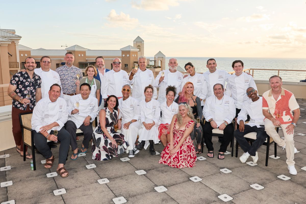 A group of chefs and guests, many in white jackets, pose and smile outdoors on a rooftop at sunset near the ocean during the Cayman Cookout. Some sit while others stand behind them, with buildings and the sea in the background. Lifestyle