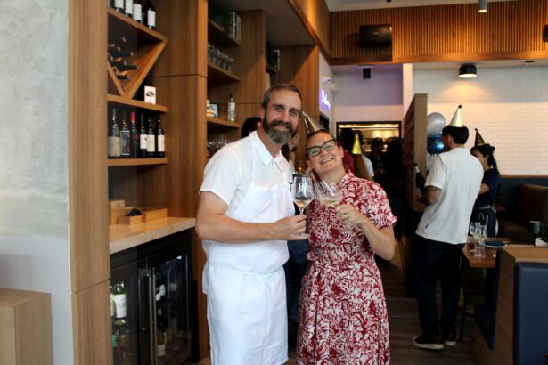 A man in a white chef’s uniform and a woman in a red patterned dress smile and toast with wine glasses in a modern restaurant, as people socialize in the background during Ukiah Celebrates First Anniversary. Lifestyle