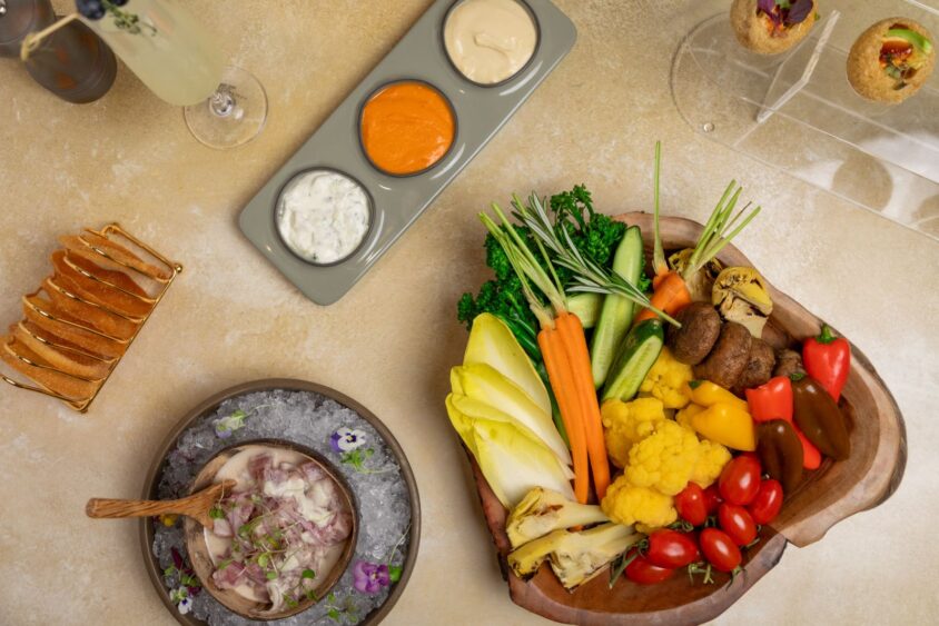 A wooden platter from Ember & Vine showcases assorted fresh vegetables, a bowl of ceviche on ice, small toast slices, three dipping sauces, and two filled pastry shells—perfect for refined Hospitality Management experiences. Lifestyle