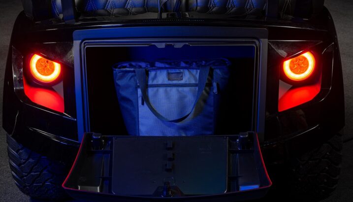 A close-up of an open car trunk with illuminated red taillights, revealing a dark blue tote bag inside under blue lighting. The car's black and sleek exterior is partially visible. Lifestyle