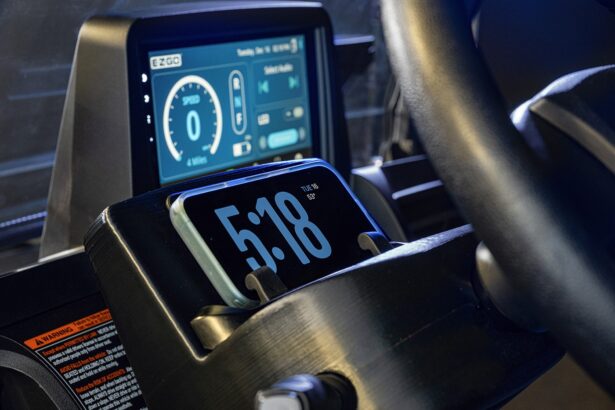Close-up of a car dashboard showing a digital display with speed and battery info. In front, a smartphone in a holder displays the time, 5:18, and the date, Tue, Sep 5. A steering wheel is partially visible on the right. Lifestyle
