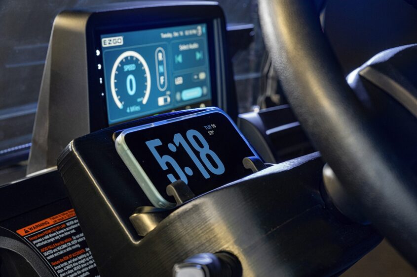 Close-up of a car dashboard showing a digital display with speed and battery info. In front, a smartphone in a holder displays the time, 5:18, and the date, Tue, Sep 5. A steering wheel is partially visible on the right. Lifestyle