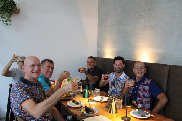 Six men sit around a wooden table at a restaurant, smiling and raising their drinks in a celebratory toast as Ukiah celebrates its first anniversary. Plates of food and festive decorations add to the cheerful, relaxed atmosphere. Lifestyle