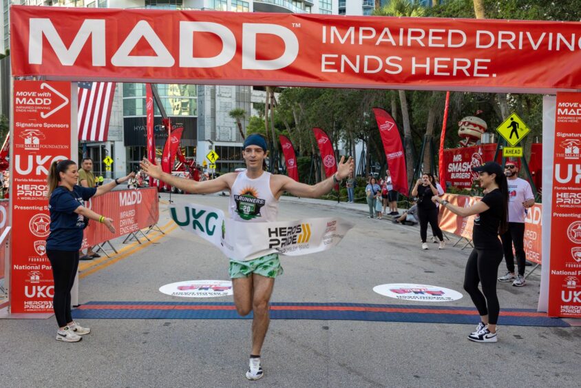 A male runner crosses the MADD Dash 5K finish line, breaking the tape and spreading his arms in triumph. An overhead banner reads “MADD IMPAIRED DRIVING ENDS HERE” as spectators cheer for the fundraising race against drunk driving. Lifestyle