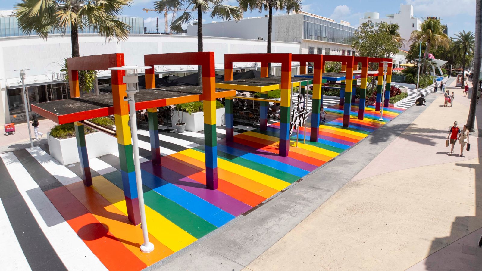 A colorful outdoor installation on Lincoln Road features rainbow-painted ground and columns with black tops, surrounded by palm trees and buildings. This vibrant Art Walk attracts people to enjoy art and love on a sunny day. Lifestyle