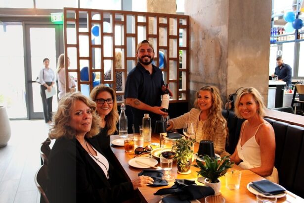 Five women are seated at a restaurant table, smiling at the camera, while a man stands beside them holding a wine bottle. The modern setting with natural light and decor hints at Ukiah Celebrates First Anniversary in style. Lifestyle