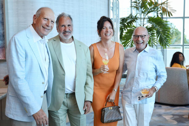 Four adults, three men in light suits and a woman in an orange dress, stand together indoors smiling at the camera during the Annual Salute to Leadership for Broward Partnership. Two hold drinks, with large windows and plants visible in the background. Lifestyle
