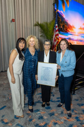 Four women in business attire stand indoors, smiling at the camera. One holds a large framed certificate. Behind them are an American flag, a potted plant, and a screen displaying a tropical beach scene at sunset. Lifestyle