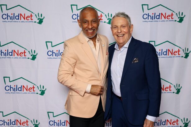 Two men smile together in front of a step-and-repeat banner featuring the ChildNet logo and green handprints at the ChildNet Hosts Inaugural Ripple Effect Gala. One sports a beige jacket, while the other is dressed in a navy blue suit. Lifestyle