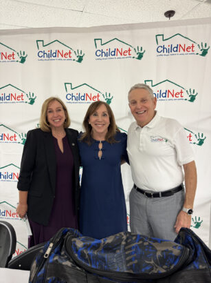 Three adults stand smiling in front of a step-and-repeat ChildNet banner. One wears a blazer, another a blue dress, and the third a white ChildNet polo shirt. A donated duffle bag is visible in the foreground, highlighting their luggage donation effort. Lifestyle