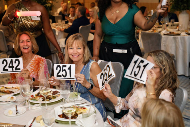 Four women seated at a dining table during the Broward Partnership Annual Leadership Event hold up numbered paddles—242, 251, 232, and 151—while smiling. Plates of food, drinks, and elegant table settings are visible in front of them. Lifestyle